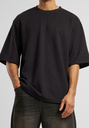 Man draagt een effen zwart oversized t-shirt met korte mouwen, met één hand in de zak en donker gewassen spijkerbroek op een neutrale achtergrond.