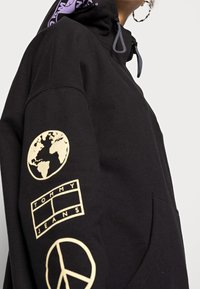 Zwarte hoodie met een capuchon met trekkoorden, bedrukt met gouden grafische ontwerpen van een wereldbol, het "TOMMY JEANS"-logo en een vredessymbool op de mouw.