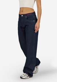 Sötétkék, laza szabású farmernadrág látható zsebekkel és a derékpánton piros hímzett márkalogóval, ezüst sportcipővel párosítva.