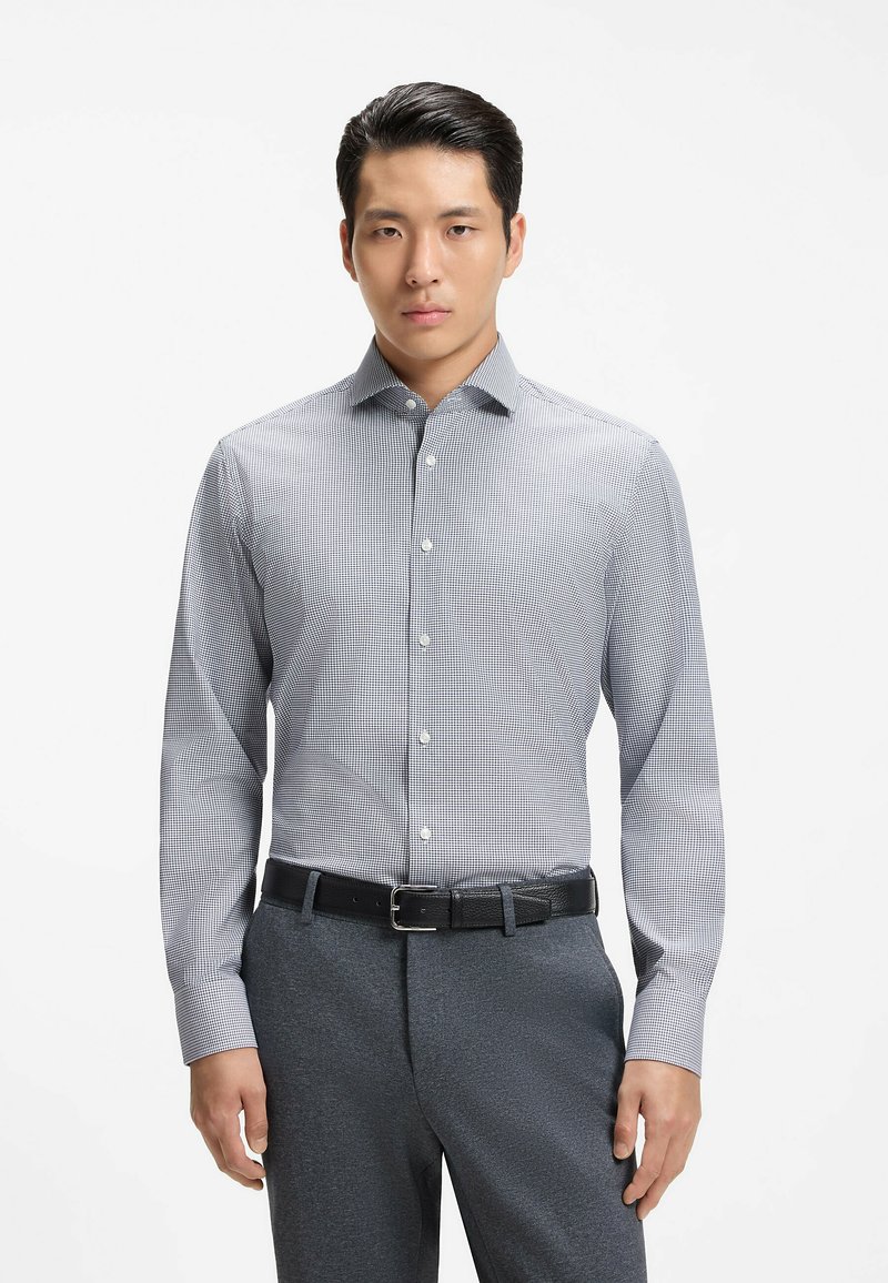 Homme portant une chemise grise à carreaux boutonnée, rentrée dans un pantalon gris foncé avec une ceinture noire, debout devant un fond blanc uni.