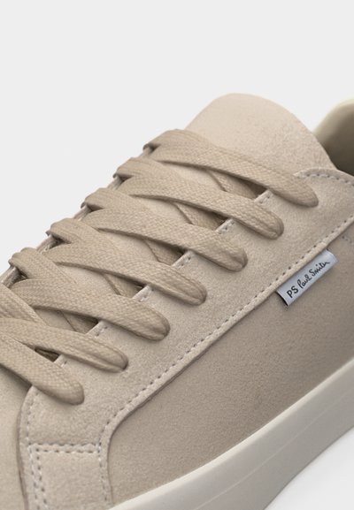 Κοντινό πλάνο μπεζ σουέντ αθλητικού παπουτσιού με ταιριαστά κορδόνια και ετικέτα "PS Paul Smith" στο πλάι κοντά στη σόλα.