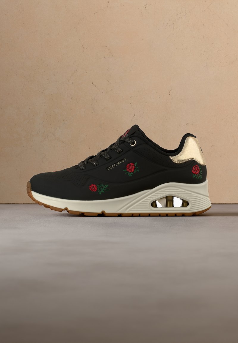 Baskets noires Skechers avec broderie de rose rouge, semelle beige avec coussin d'air, lacets noirs et détail métallique doré au talon.