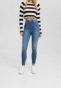 Mujer vestida con jeans skinny azul de tiro alto, zapatillas blancas y un suéter corto a rayas blancas y negras sobre un fondo gris.
