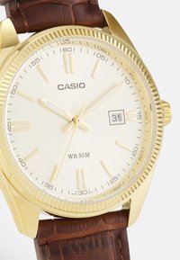 Gold-tone Casio watch with a round white dial, brown textured leather strap, date window at 3 o'clock, and luminous hour and minute hands.
