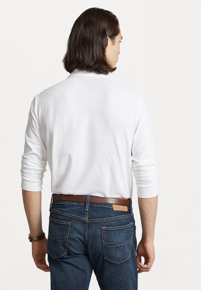 Witte lange mouwen shirt met ribbelstructuur, gecombineerd met donkerblauwe jeans en een brown riem. Zichtbare achternaden en zakdetails.