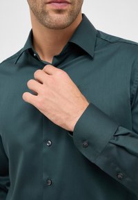 Camicia da uomo verde scuro con texture liscia, collo a punta e polsini abbottonati. Presenta bottoni neri e un design aderente.