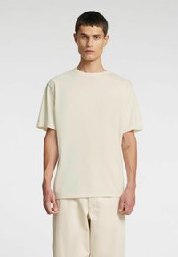 Beżowy bawełniany t-shirt z krótkim rękawem, luźnym krojem i okrągłym dekoltem. Noszony z dopasowanymi beżowymi spodniami; gładka powierzchnia, bez wzorów.