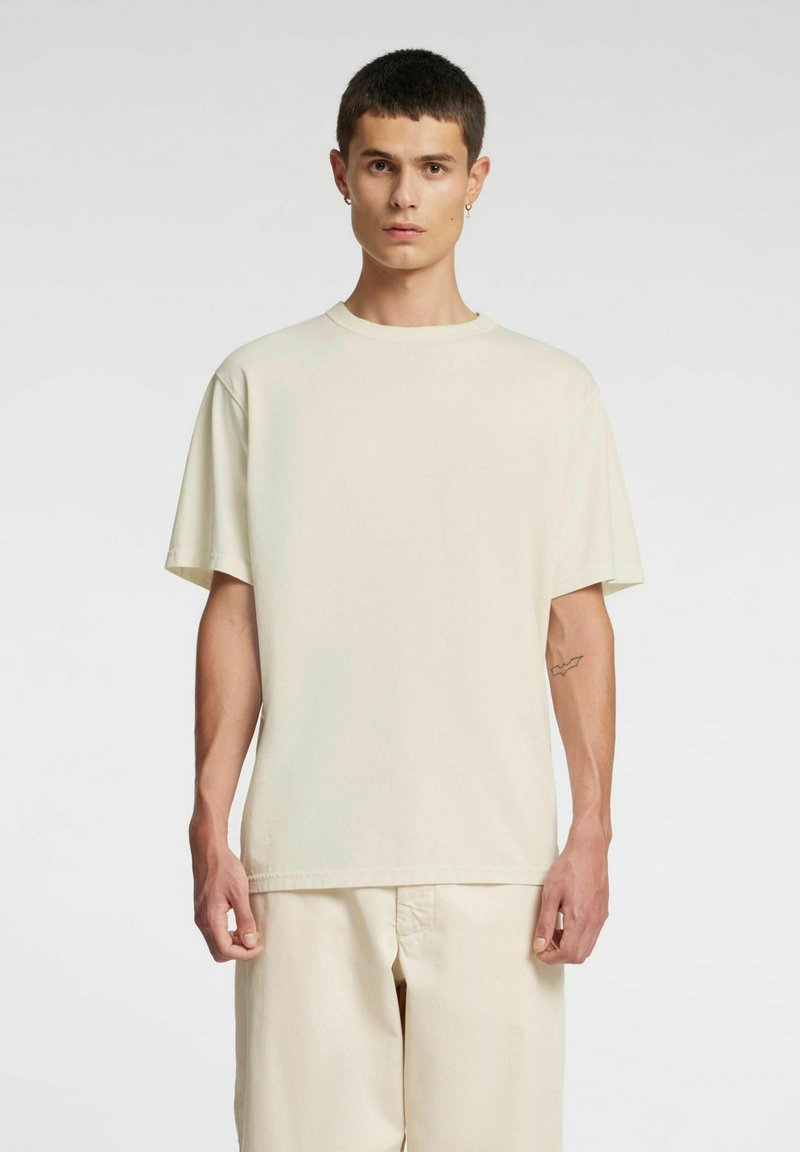 Beżowy bawełniany t-shirt z krótkim rękawem, luźnym krojem i okrągłym dekoltem. Noszony z dopasowanymi beżowymi spodniami; gładka powierzchnia, bez wzorów.