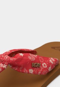 Καφέ σανδάλι με κόκκινο λουρί υφάσματος με σχέδιο, δεμένο σε κόμπο στο πλάι, και μεταλλικό λογότυπο "ROXY" στην κορυφή του λουριού.