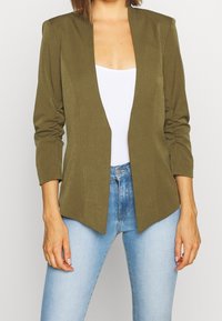 Olive green blazer with a notched collar, open front design, and ruched sleeves. Worn over a white top and paired with light blue jeans.