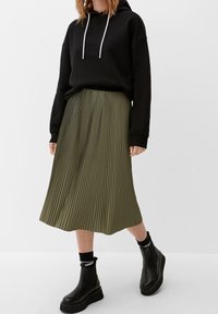 Sudadera negra con cordones blancos, combinada con una falda plisada verde. La modelo lleva botas gruesas negras y calcetas, mostrando un estilo en capas.