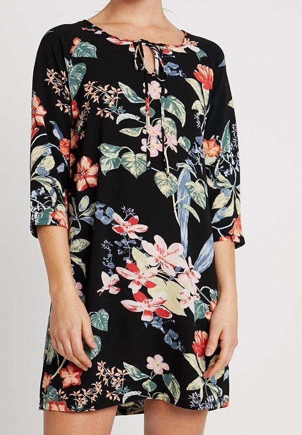 Femme portant une robe noire à motifs floraux multicolores vifs et manches trois quarts, avec un petit lien au niveau de l'encolure.