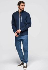 Marinblå zip-up jacka med mandarin-krage, texturerat tyg och två dragkedjefickor; kombinerad med ljusblå jeans och bruna sneakers.