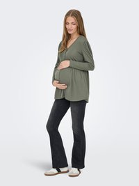 Zwangere vrouw die een olijfgroene blouse, zwarte jeans en witte sneakers draagt, zachtjes haar buik vasthoudend en naar beneden kijkend.