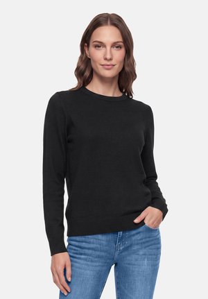 Femme aux cheveux bruns mi-longs, portant un pull noir à manches longues et un jean bleu, debout avec une main dans la poche.