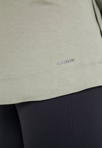 T-shirt de sport vert clair avec une texture lisse, arborant le logo "AEROREADY" à l'ourlet, assorti à des leggings noirs.