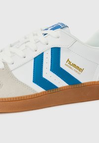 White athletic shoe featuring blue accents and a beige suede toe cap, with a textured gum rubber sole and perforated detailing.
