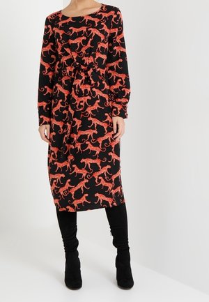 Schwarzes Kleid mit rotem Leopardenmuster, lange Ärmel, tailliert, fällt unter das Knie. Kombiniert mit schwarzen hohen Stiefeln.