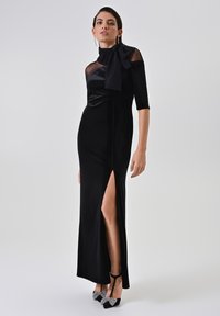 Zwart fluwelen avondjurk met een hoge split en doorzichtige mouwaccenten. Heeft een strikdetail bij de hals en een slank, aansluitend ontwerp.