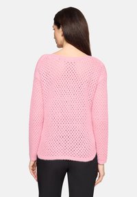 Femme aux longs cheveux bruns portant un pull en tricot ouvert rose et un pantalon noir, vue de dos sur un fond uni.