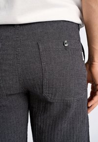 Grijze herringbone-patroon broek met een achterzak met knoopsluiting, gemaakt van een gestructureerde stof. Hand nabij de zak gepositioneerd.