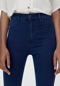 Jean taille haute bleu foncé à texture lisse, avec une fermeture à boutons à l'avant et un design à cinq poches, présenté sur un fond clair.