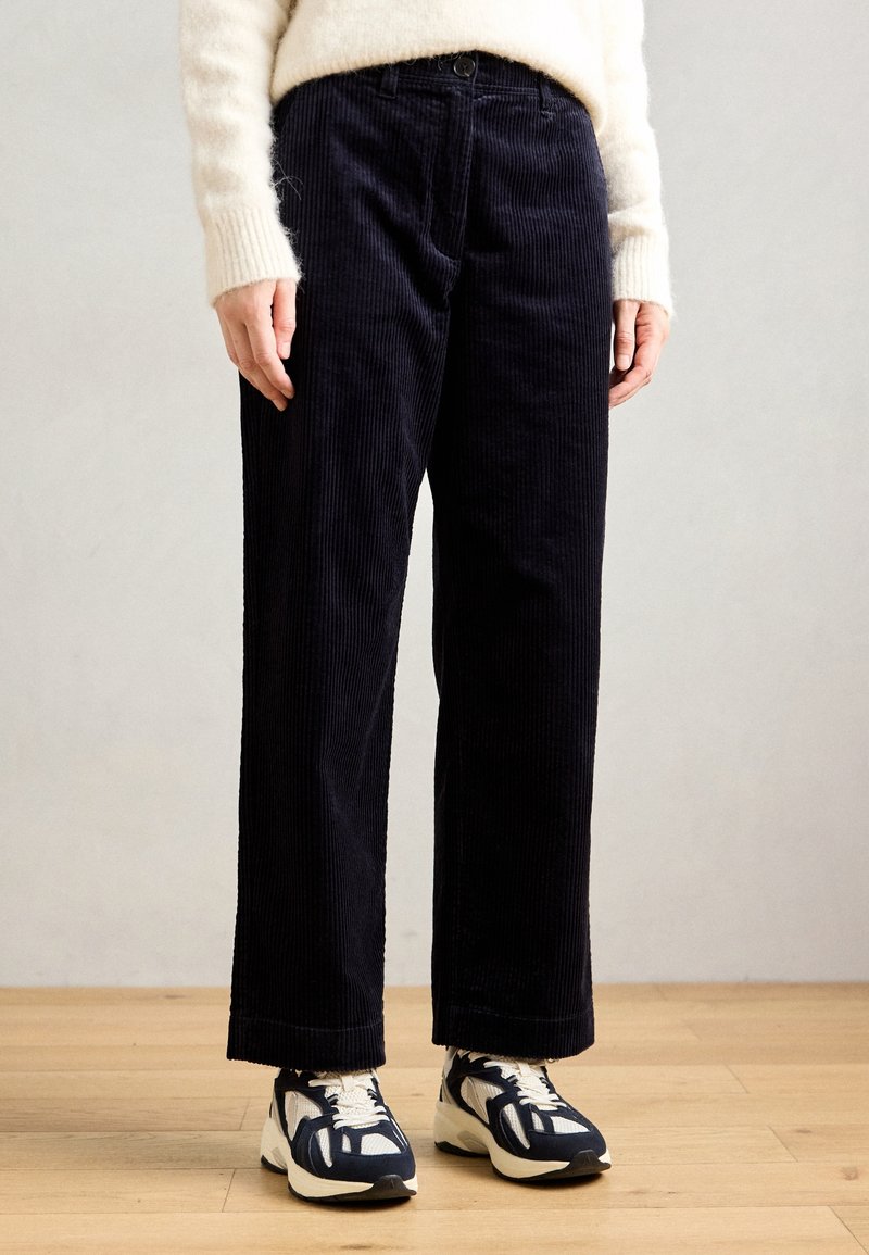 Personne portant un pantalon large en velours côtelé noir, des baskets blanches et bleu marine, et un pull crème, debout sur un parquet en bois.