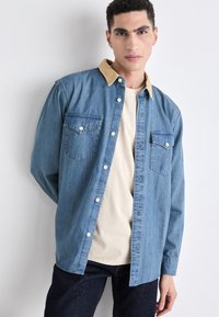 Denim shirt with light blue fabric, tan collar, button-up front, two chest pockets, and long sleeves, worn over a cream-colored t-shirt.