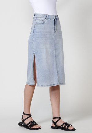 Hellblaue Jeans-Midi-Rock mit einem Schlitz vorne, schrägem Saum und Fünf-Taschen-Design. Kombiniert mit schwarzen, schräg geschnittenen Sandalen.