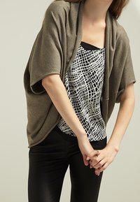 Warstwowy sweter w oliwkowej zieleni na czarno-białym wzorzystym topie; modelka ma na sobie czarne dopasowane spodnie o gładkiej fakturze i luźnym kroju.