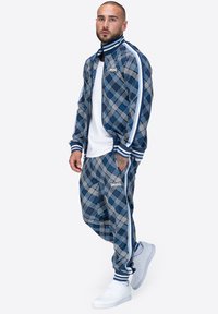 Mėlynos struktūrinės vilnos sportinis kostiumas su užtrauktuku, turintis baltas detales ir atitinkančias kelnės, derinamas su baltomis sportinėmis bateliais.