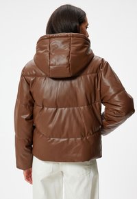 Brun faux læder pufferjakke med hætte, der har et quiltet design og elastiske manchetter. Bagudsigten understreger dens afrundede, puffy form.