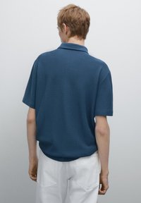 Námořnická modrá polo košile s krátkým rukávem, texturovaná látka, volný střih a klasický límeček, v kombinaci s bílými kalhotami.