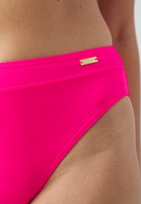 Parte de baixo de biquíni rosa, feita de material liso e elástico. Apresenta uma etiqueta lateral com letras douradas e um design minimalista com costuras planas.