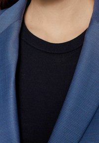 Blazer bleu en tissu texturé, associé à un t-shirt col rond bleu marine foncé. Accent sur le col et les détails des coutures aux épaules.