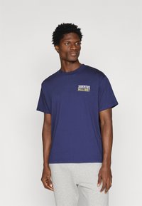 T-shirt de algodão azul-marinho com mangas curtas, apresenta o texto "JUVENTUS MILLE897" em branco e amarelo no peito. Design minimalista.