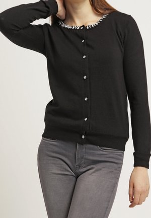 Frau trägt einen schwarzen, geknöpften Cardigan mit juwelenverziertem Ausschnitt und graue, taillierte Hose, ein Arm ist erhoben und hinter den Kopf gebogen.