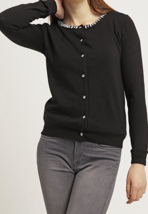 Femme portant un cardigan noir boutonné avec un col orné de bijoux et un pantalon gris ajusté, un bras levé et plié derrière la tête.
