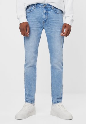 Mężczyzna ubrany w jasnoniebieskie obcisłe dżinsy, białe trampki i biały długi rękaw, stojący na tle jednolitego jasnego tła.