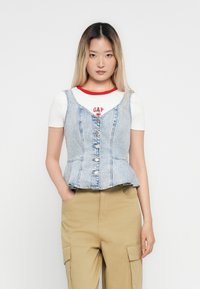 Denim peplum top in lichtblauwe kleur, met een knoopsluiting aan de voorkant en verstelbare banden over een witte blouse met rode accenten.