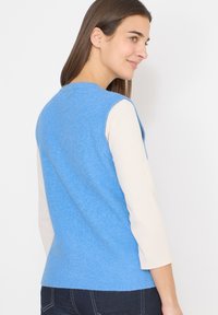Femme aux longs cheveux bruns portant un gilet en tricot bleu sans manches sur une chemise crème à manches longues et un jean en denim foncé, faisant face à droite.