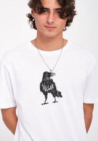 T-shirt en coton blanc avec un graphisme noir d'un corbeau intégré au design avec le texte "VOLCOM". Manches courtes et coupe décontractée.