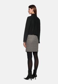 Schwarze, taillierte Jacke mit hohem Kragen und gerafftem Bund, kombiniert mit einem gemusterten grau-schwarzen Rock, schwarzen Strumpfhosen und Stiefeletten.