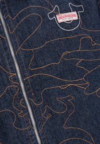 Spijkerjasje met een donkerblauwe basis en contrasterende oranje stiksels. Voorzien van een ritssluiting aan de voorkant en een True Religion-logo patch.