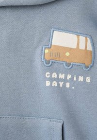 Světle modrá mikina s kapucí a měkkou texturou. Obsahuje vyšitý grafický prvek s vanem v béžové a bílé barvě a text "CAMPING DAYS" nad přední kapsou.