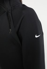 Černá sportovní bunda s texturovanou tkaninou, částečným zipem a bílým logem Nike na levém rameni.