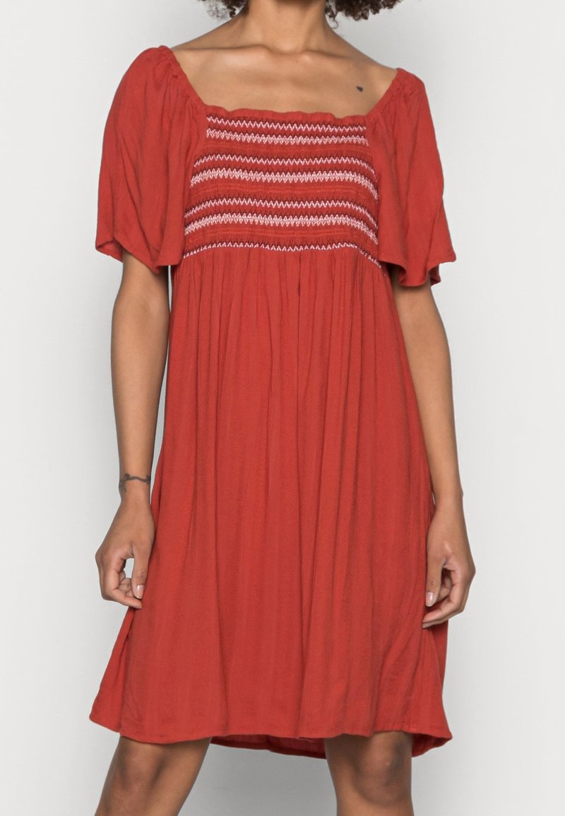 Robe rouge à manches courtes avec corsage smocké élastique présentant des coutures blanches en chevrons, portée par une personne les mains détendues le long du corps.