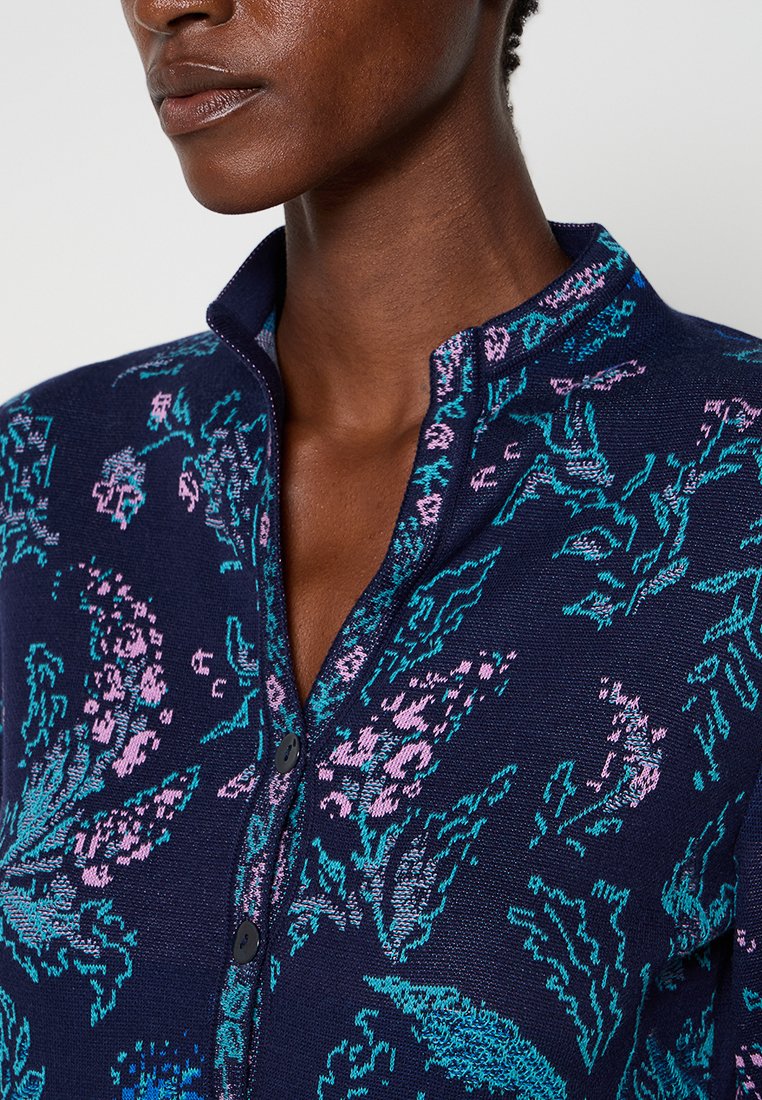 Chemise tricotée bleu marine avec un motif floral en turquoise et rose. Dispose d'un col mandarin et d'une fermeture à boutons. Texture lisse.