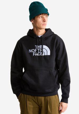 Młody mężczyzna w czarnej bluzie z kapturem The North Face, zielonej dzianinowej czapce i brązowych spodniach, patrzący na bok z rękami w kieszeniach.