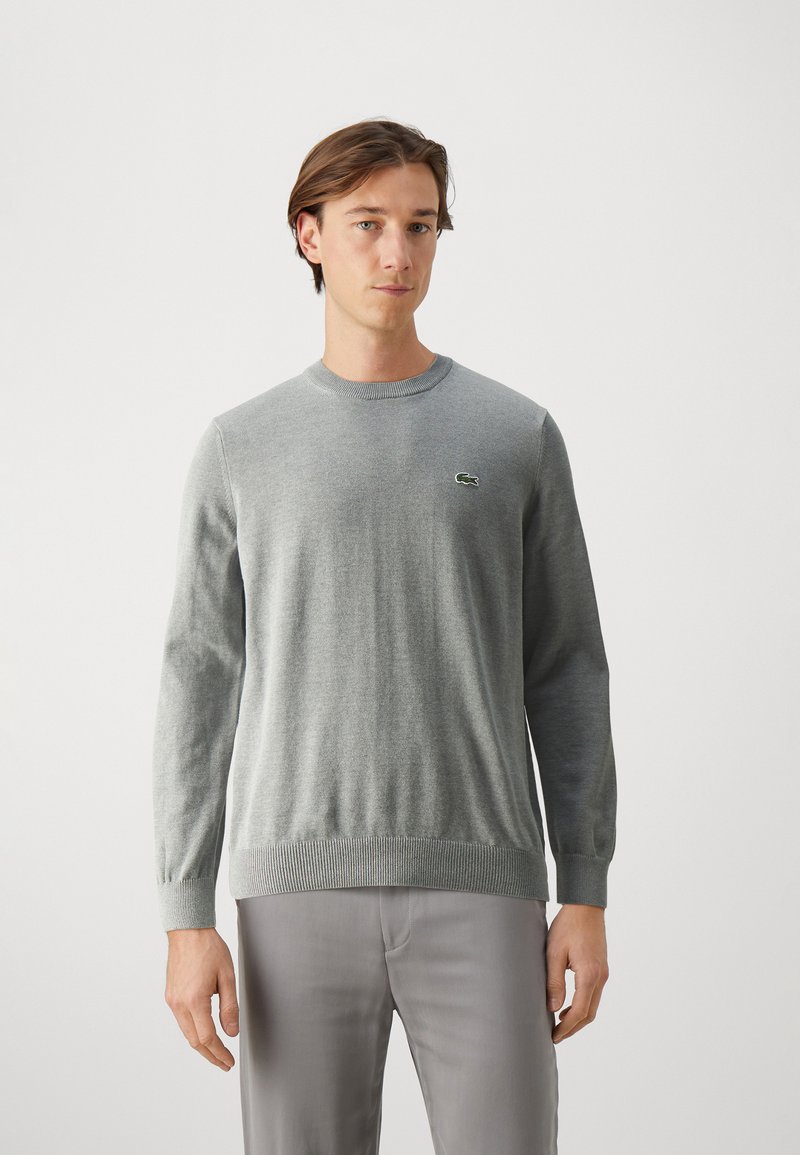 Grijze longsleeve trui met ronde hals, geribbelde manchetten en zoom, met een klein groen Lacoste-logo op de borst. Gladde brei-textuur.